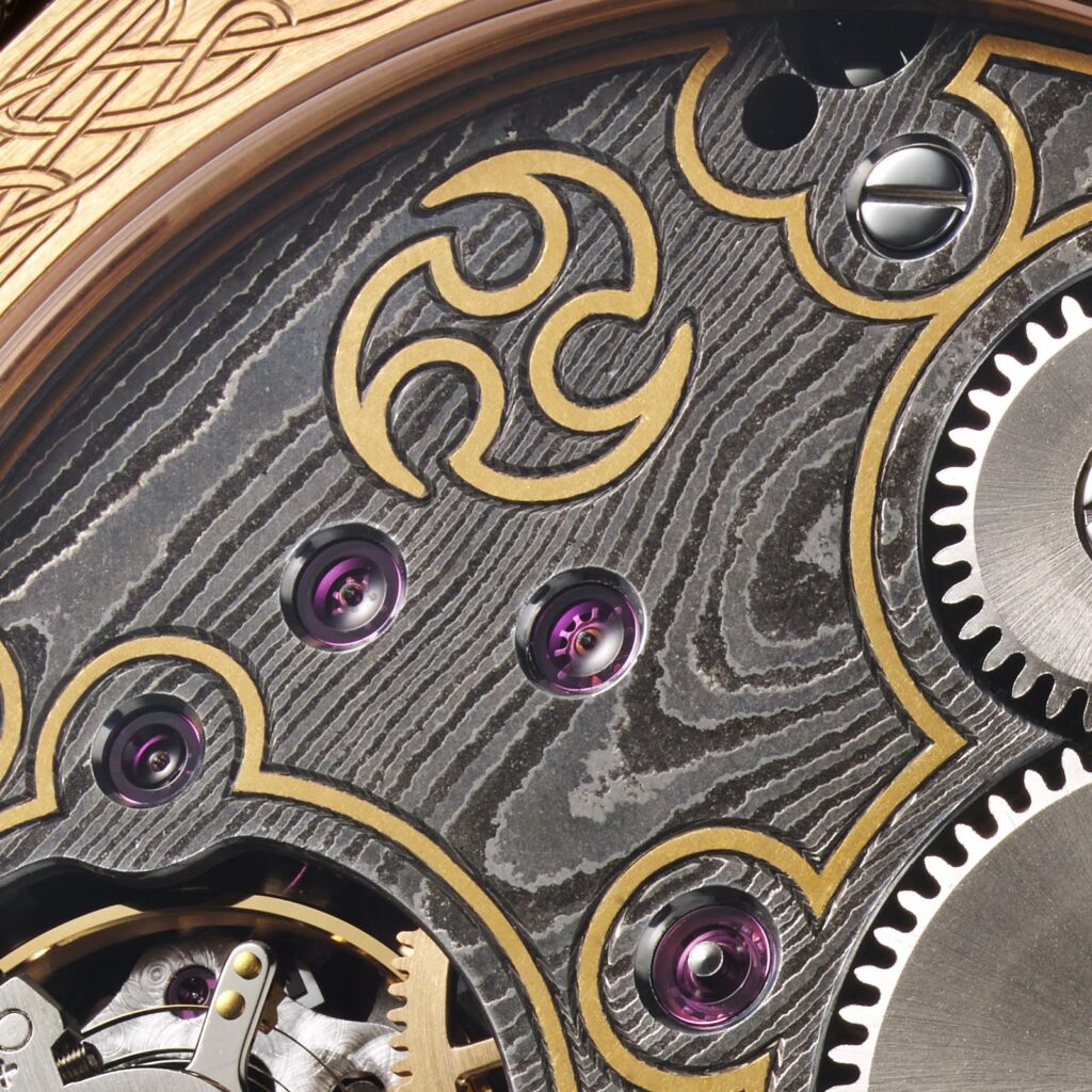 Detail of a GoS Skadi movement. The layered damascus pattern is brought up by acid etching after forging; the applied gold element draws on Norse iconography.