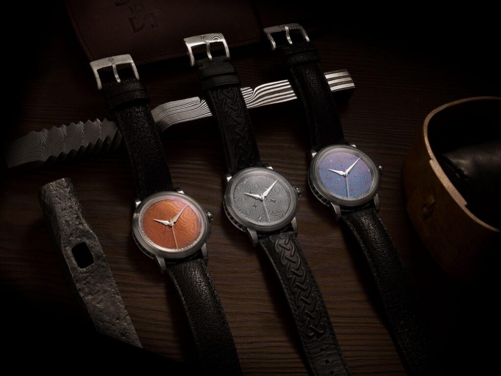 Three watches from the GoS Fullerö collection, photographed alongside a hand-forged damascus blade. The same metalwork tradition that shapes the sword produces the dials.